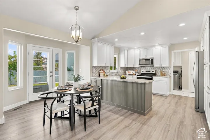 Open-concept kitchen and dining area featuring white cabinetry, stainless steel appliances, a green-painted island with waterfall countertop, subway tile backsplash, wood-finish flooring, and expansive glass doors overlooking waterfront scenery