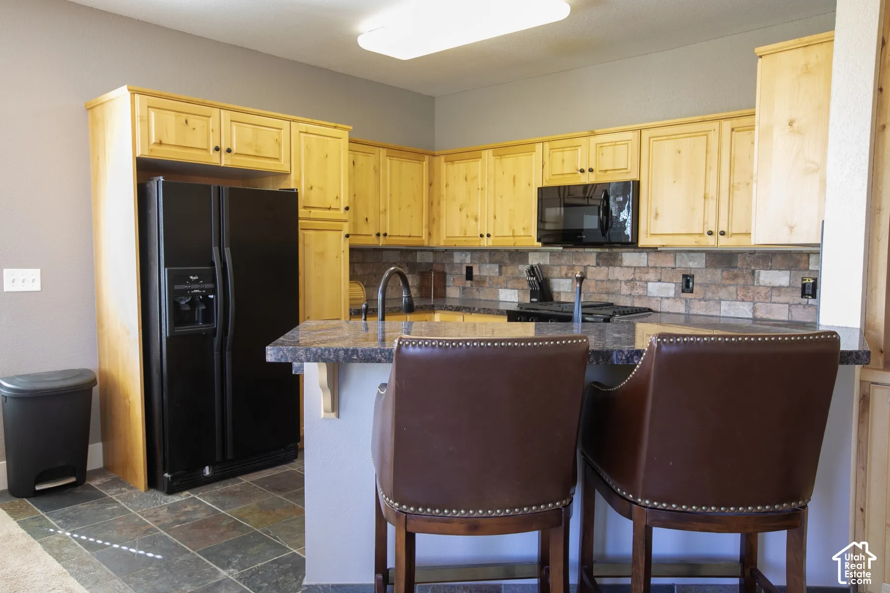 Light-toned wood cabinetry with dark hardware, dark stone-finish countertops, black built-in appliances, and a brick-patterned backsplash