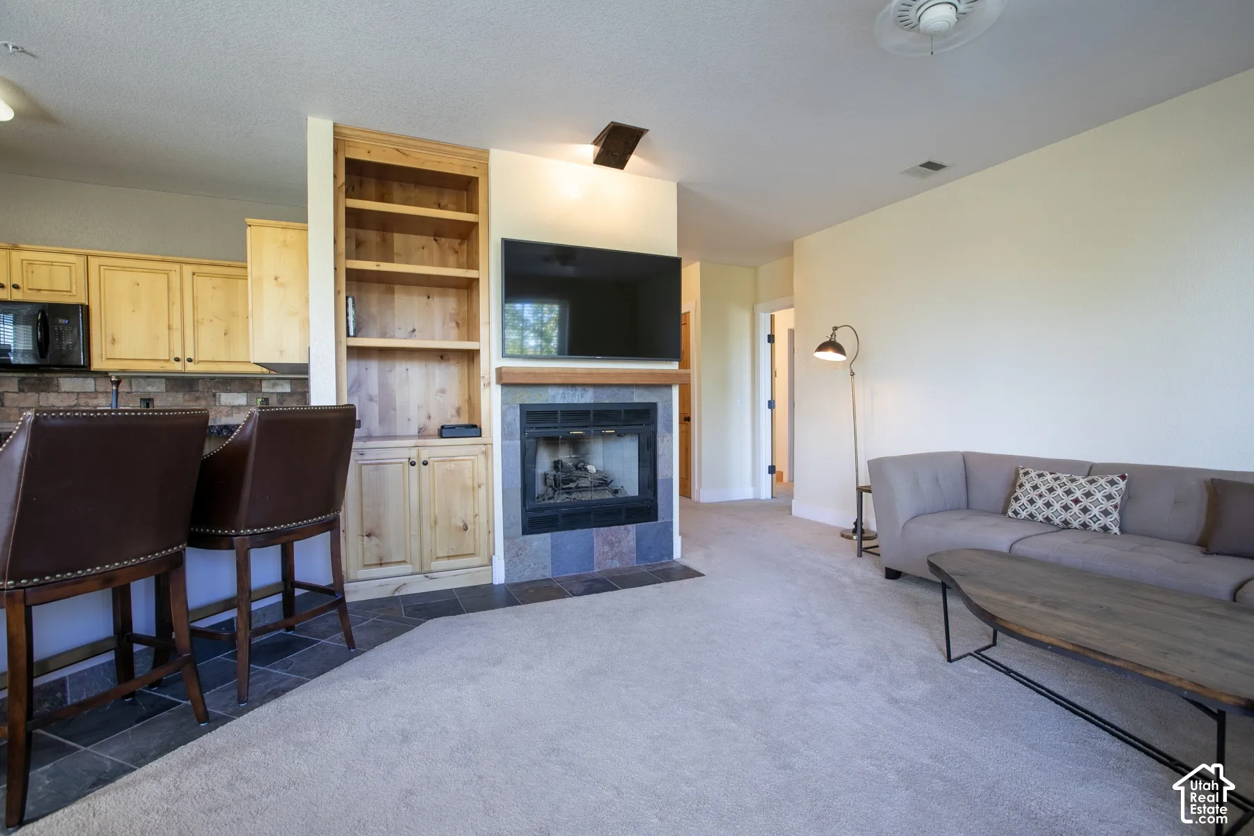 Spacious open-concept living area featuring wall-mounted flat-screen entertainment center with built-in shelving, gas fireplace with blue tile surround, neutral carpet flooring, and abundant natural lighting from multiple windows