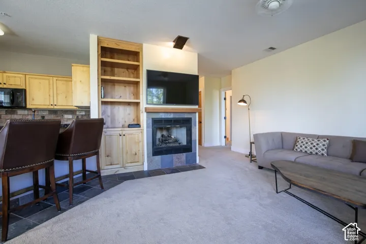 Spacious open-concept living area featuring wall-mounted flat-screen entertainment center with built-in shelving, gas fireplace with blue tile surround, neutral carpet flooring, and abundant natural lighting from multiple windows
