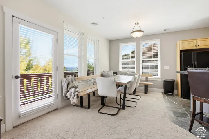 Bright and airy dining area showcasing multiple glass doors and windows with plantation shutters, neutral carpeting, a pendant light fixture, and seamless sightlines to the kitchen and adjacent spaces with stainless steel appliances visible in the backgro