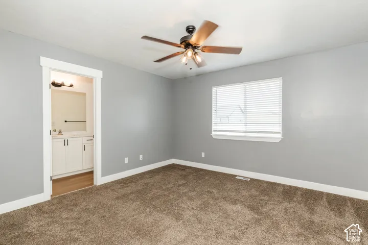 Plush carpeted flooring, a ceiling fan with integrated lighting, a window with horizontal blinds, and an attached bathroom featuring a vanity with a countertop and undermount sink