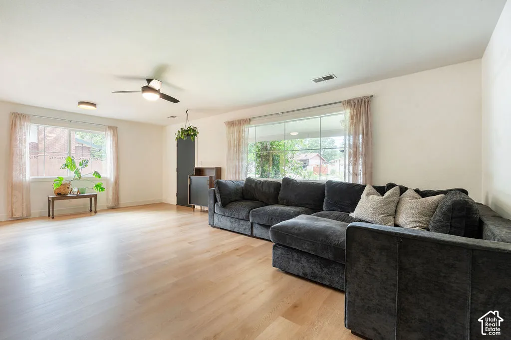 Spacious living room showcasing wood-finish flooring, abundant natural light from multiple large windows with sheer drapery, a ceiling fan, and an open floor plan ideal for entertaining