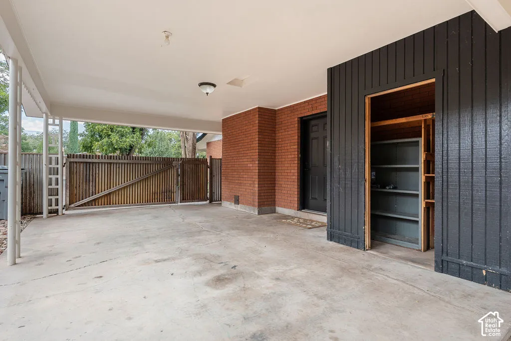 Covered concrete carport featuring a secure wooden gate, brick exterior, and built-in storage shelving