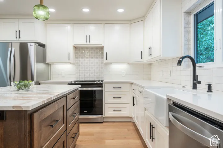 White shaker cabinetry with matte black hardware, wood-finish flooring, stainless steel appliances, a farmhouse sink with a gooseneck faucet, and an island featuring a light-toned countertop