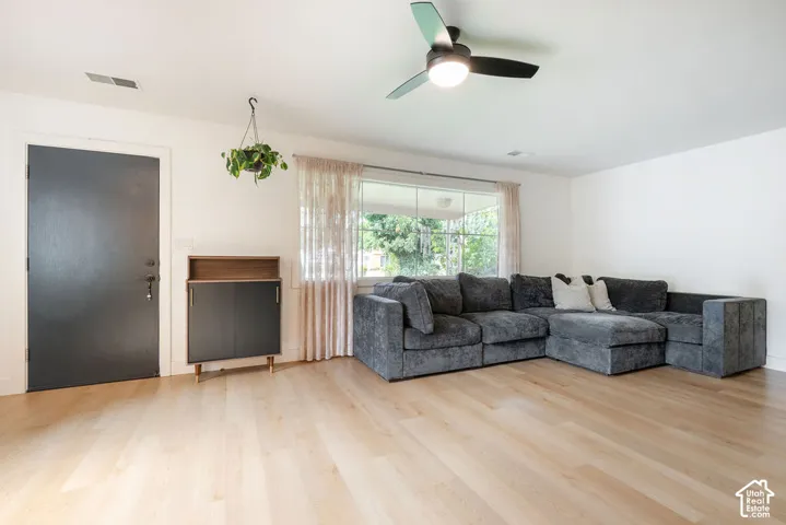 Spacious living room with light wood-finish flooring, expansive windows with natural light, ceiling fan with integrated lighting, and a dark entry door