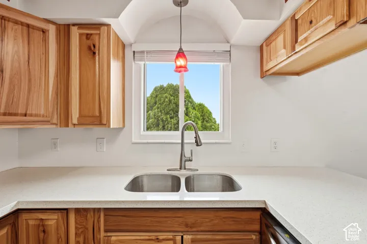Kitchen featuring natural wood cabinetry, solid-surface countertops, dual-basin stainless steel sink with a high-arch pull-down faucet, and a decorative pendant light fixture beneath a vaulted ceiling detail