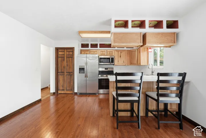 Kitchen featuring wood-finish flooring, stainless steel appliances, light wood cabinetry, white countertops, and a breakfast bar