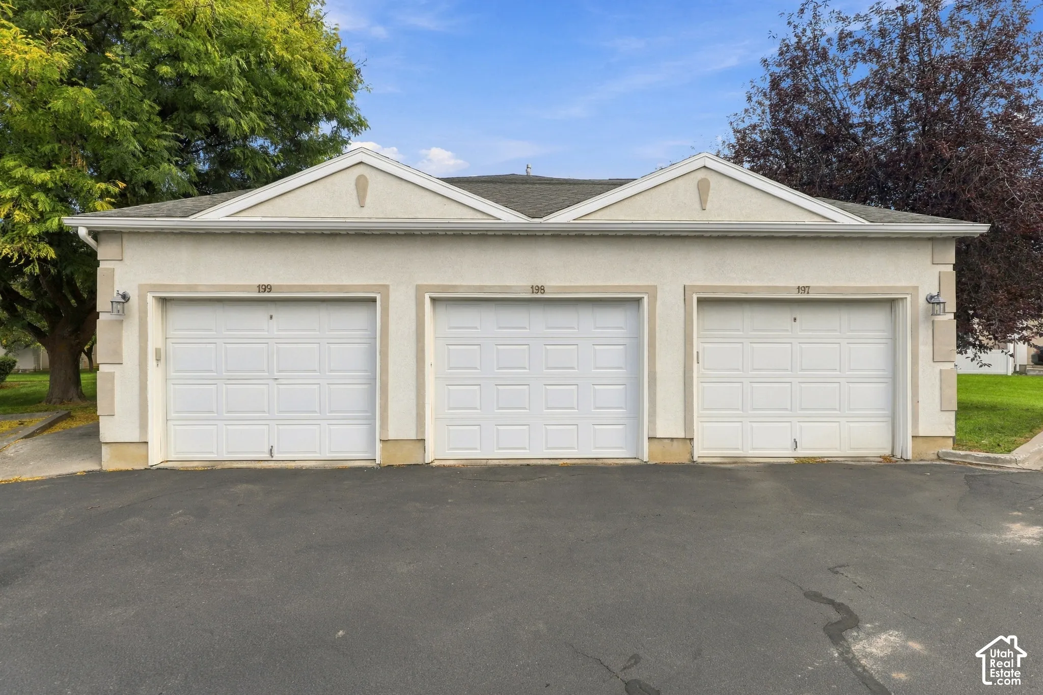 Three-bay garage featuring white paneled doors, neutral stucco exterior with brick foundation accents, and pitched rooflines with gable detailing