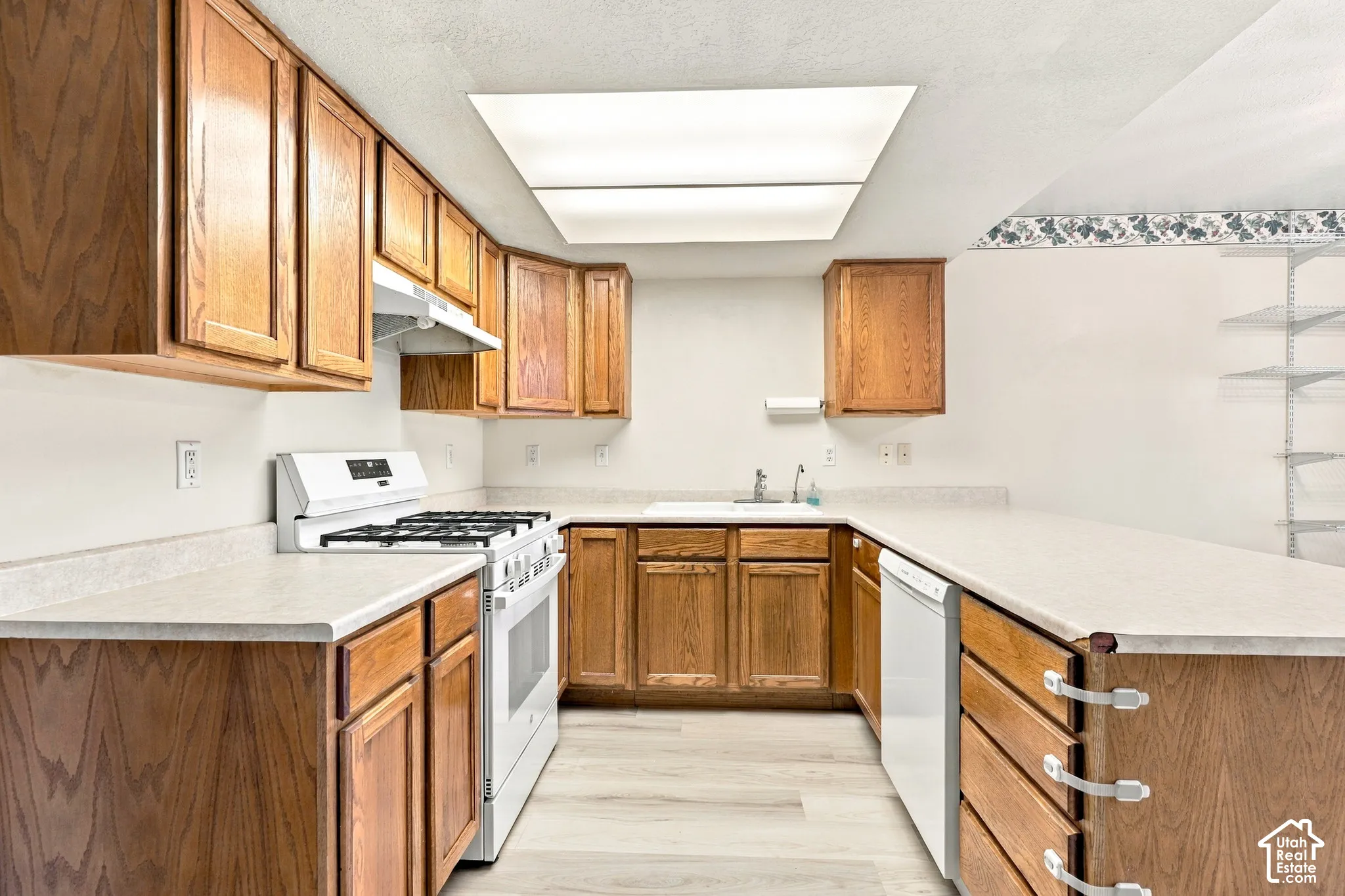 Galley kitchen featuring warm wood cabinetry, white countertops, white subway tile backsplash, stainless steel gas range, and abundant natural light from a skylight above