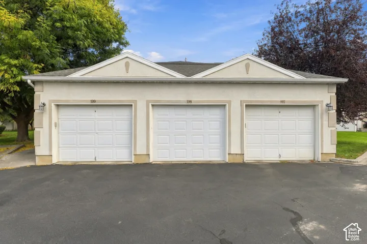 Three-bay garage featuring white paneled doors, neutral stucco exterior with brick foundation accents, and pitched rooflines with gable detailing