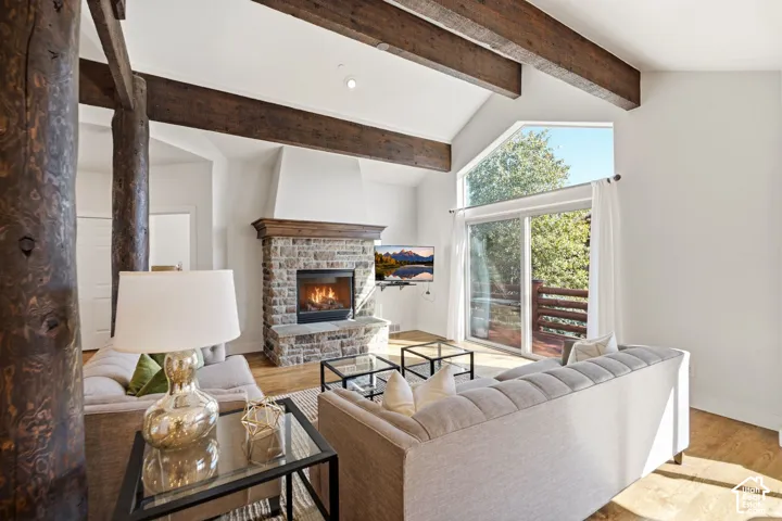 Vaulted ceilings with exposed wood beams, a stone-clad fireplace with wood mantel, hardwood-finish flooring, and large sliding glass doors leading to an outdoor deck