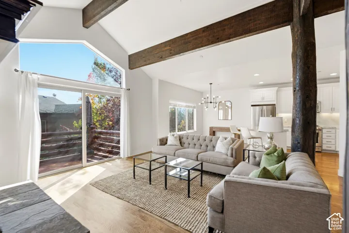 Vaulted ceilings with exposed wood beams, bright sliding glass doors, upper transom windows, wood-finish flooring, and a modern chandelier