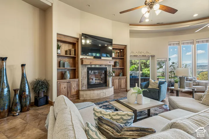 Living space featuring a stone-clad fireplace, integrated wooden shelving, a ceiling fan, and large windows with a prominent exterior landscape