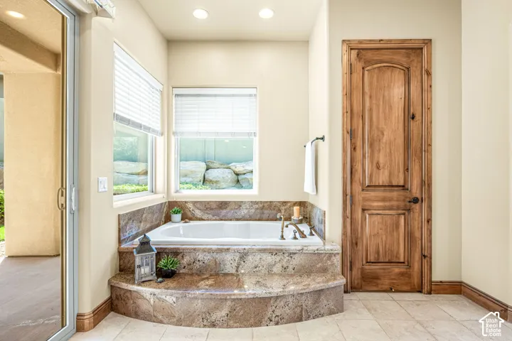 Built-in soaking tub with a stone surround, dual windows with blinds, wood-paneled door, and recessed lighting