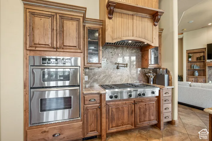 Gourmet kitchen featuring stainless steel double ovens, a built-in range with a pot filler, wood cabinetry, a granite backsplash, and tile flooring