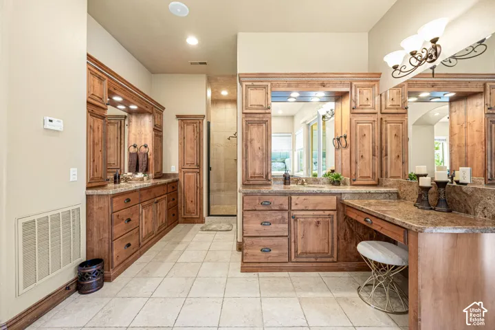 Expansive vanity area with dual sinks, wood-finish cabinetry, stone-finish countertops, tiled flooring, and a glass-enclosed shower
