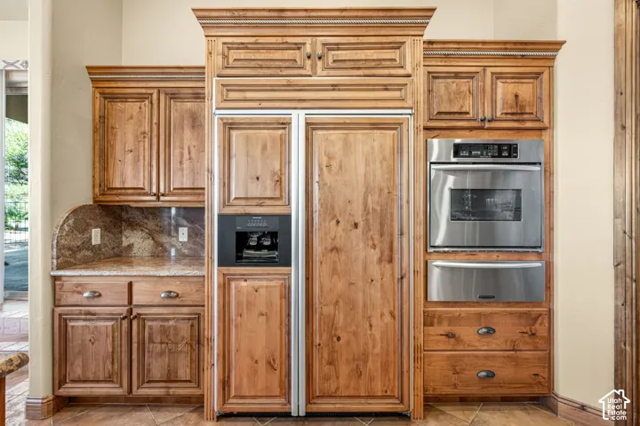 Built-in stainless steel oven, integrated paneled refrigerator, wood cabinetry with crown molding, natural stone countertop, and tile flooring