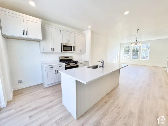Kitchen featuring an island with an integrated sink, white shaker cabinetry, stainless steel appliances, and light wood-finish flooring