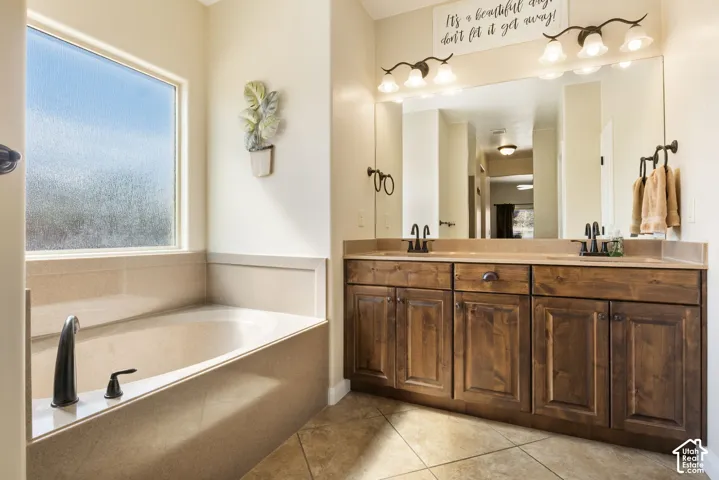 Built-in soaking tub with a frosted window, dual vanity with wood-finish cabinetry, and a large wall-mounted mirror with overhead lighting
