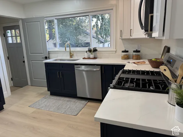 White countertops, dark blue base cabinetry, light wood-finish flooring, stainless steel appliances, and a gold-finish faucet