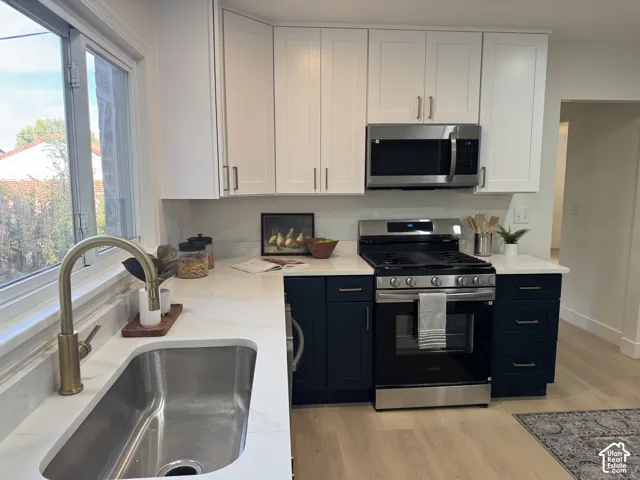 Kitchen featuring white upper cabinetry, navy blue base cabinetry, stainless steel appliances, a stainless steel sink with a gold-finish faucet, and light wood-finish flooring