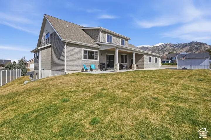 Gable roofline, shingle siding, covered patio with stone columns, concrete patio, and a large grassy yard