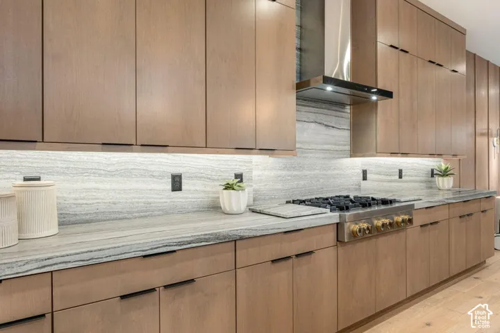 Wood-finish cabinetry with linear hardware, light-toned stone countertops, integrated gas range, stainless steel range hood, and under-cabinet lighting
