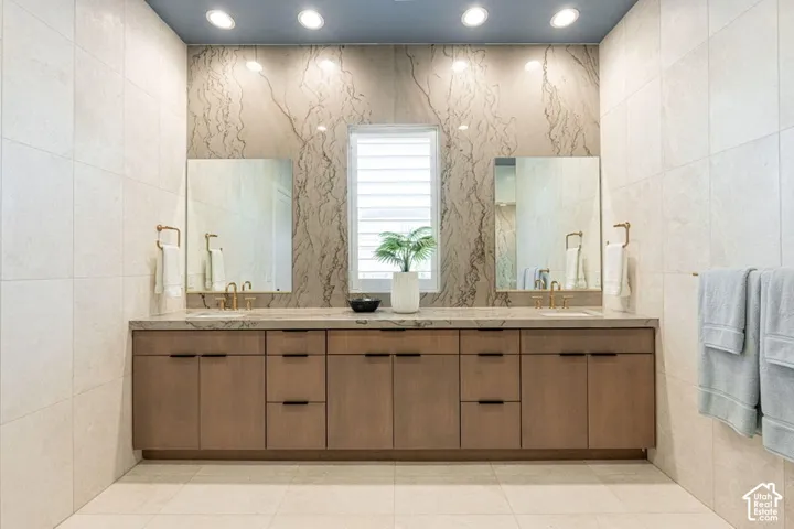 Dual vanity with integrated sinks, wood-finish cabinetry, neutral tiled walls, and recessed lighting