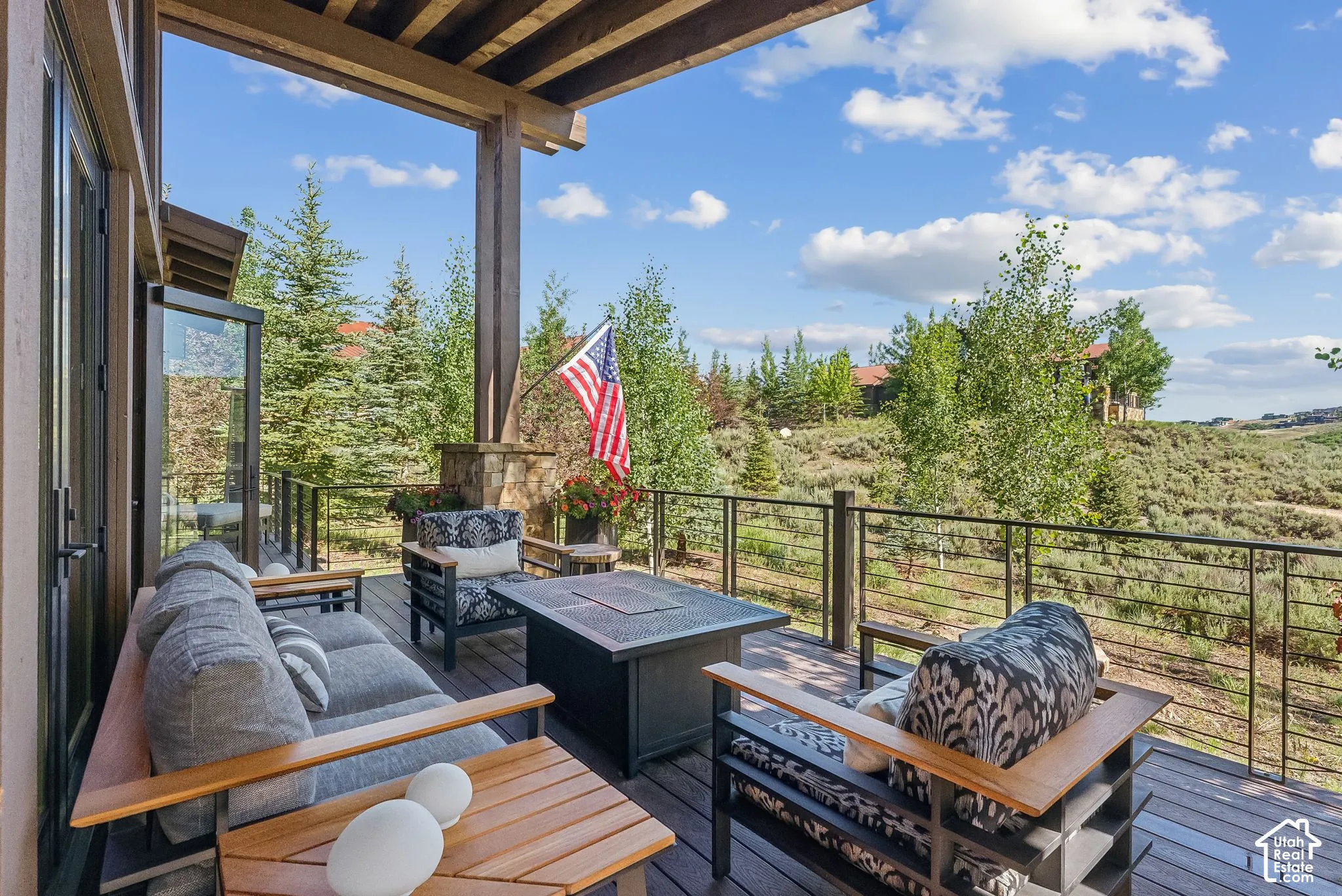 Expansive covered deck with wood-finish flooring and metal cable railing overlooking landscaped grounds with mature evergreen and deciduous trees