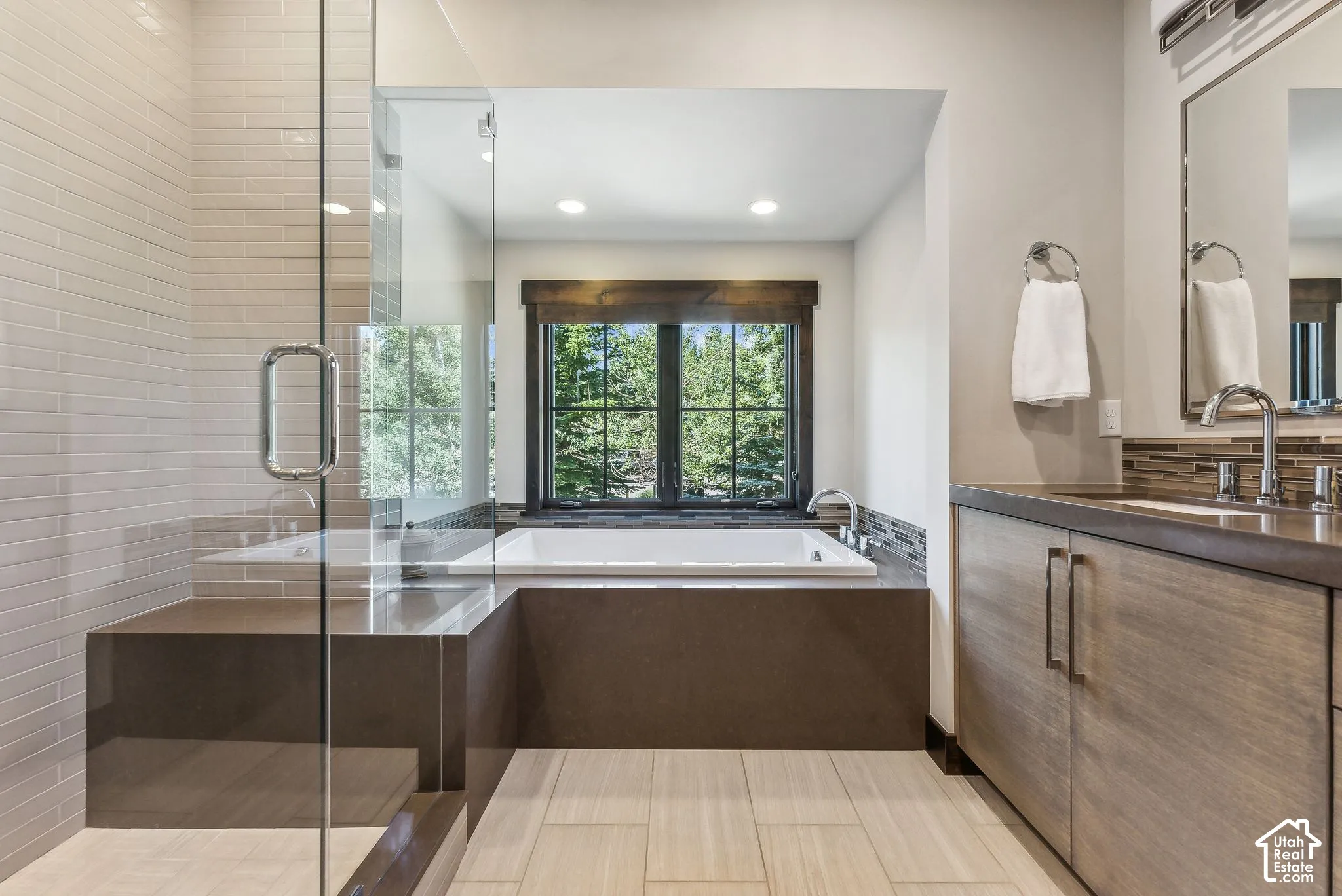 Built-in soaking tub with surrounding tile, glass-enclosed shower with tile surround, wood-finish vanity with countertop, and large framed mirror