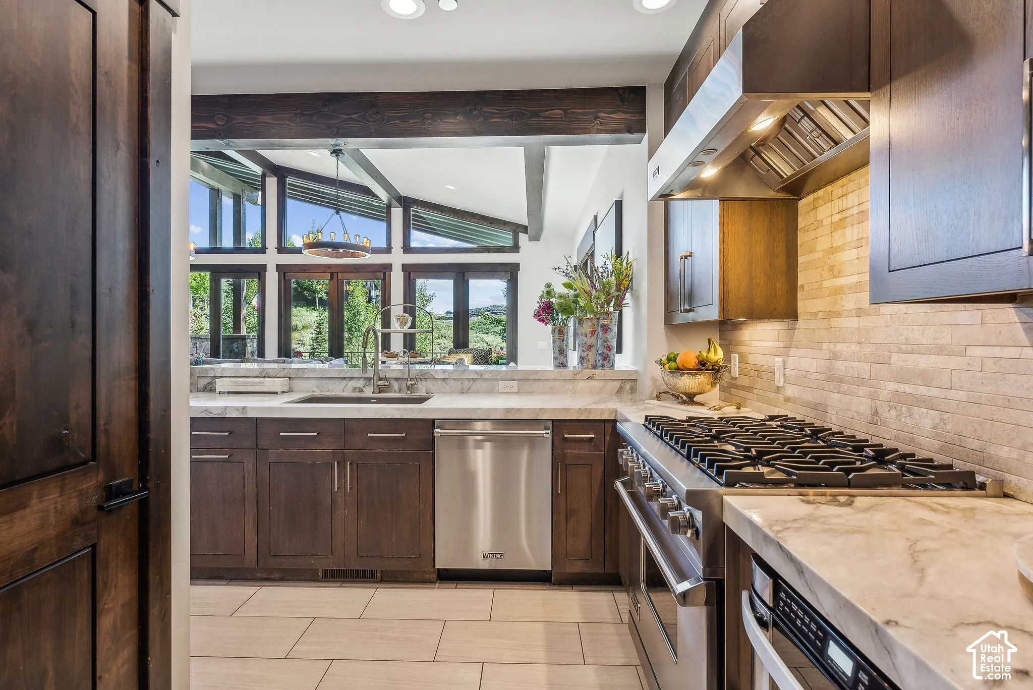 Wood-finish cabinetry, stainless steel appliances, stone slab countertops, tile backsplash, and large windows with exposed beam accents