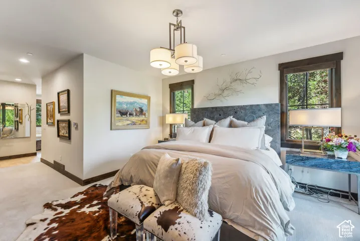 Master bedroom suite featuring a tufted upholstered headboard, layered bedding in neutral tones, chrome pendant lighting fixture, wood-finish flooring with cowhide accent rug, and French doors opening to outdoor spaces with garden views