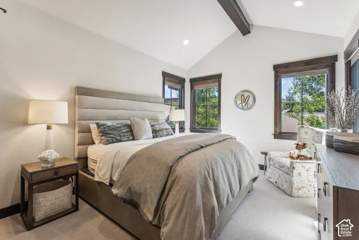 Primary bedroom suite featuring a horizontal-slat upholstered headboard with integrated nightstands, layered bedding in neutral tones, vaulted ceiling with exposed beam detailing, recessed lighting, large multi-pane windows with dark wood frames offering