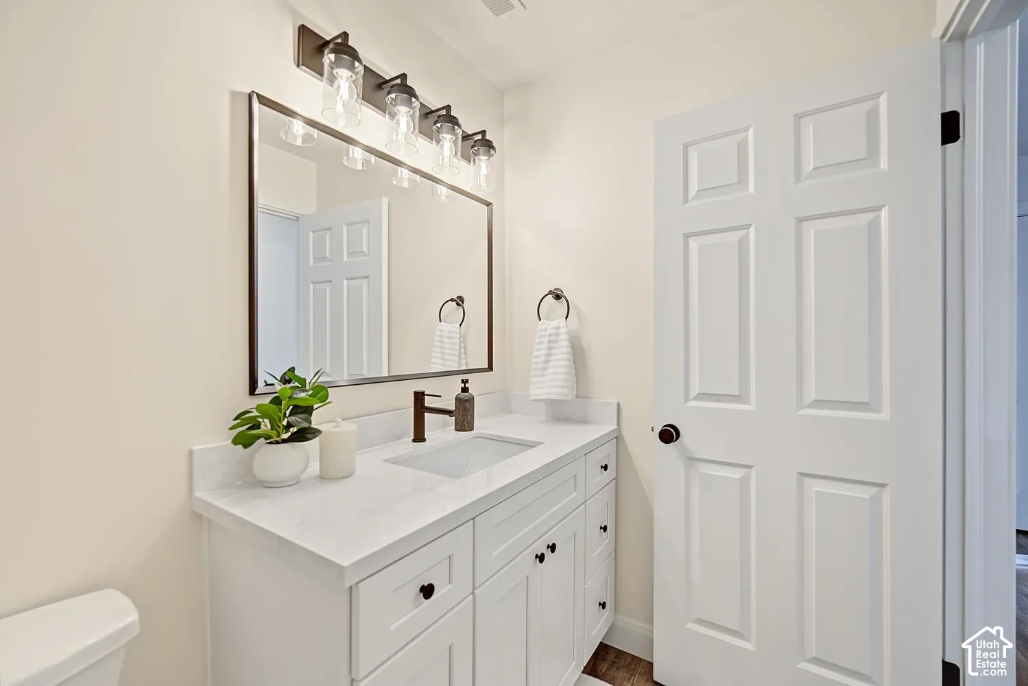 White vanity with black hardware, white countertop with integrated sink, rectangular mirror with dark frame, four-light vanity fixture with clear glass shades, and dark bronze faucet
