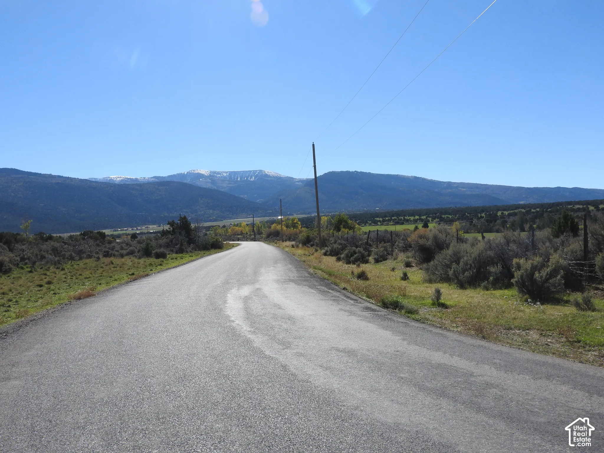 Asphalt paved road flanked by native scrub brush, rolling terrain, and a prominent mountain range backdrop