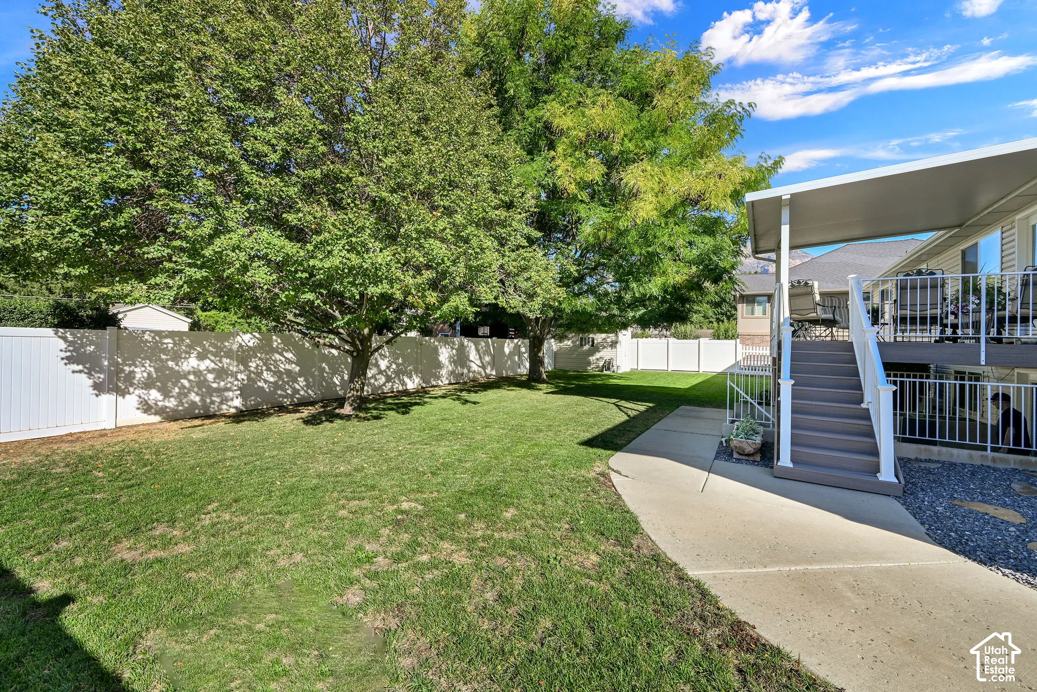 Expansive backyard with a manicured lawn, mature trees, white privacy fencing, a covered deck, and exterior stairs with white railings