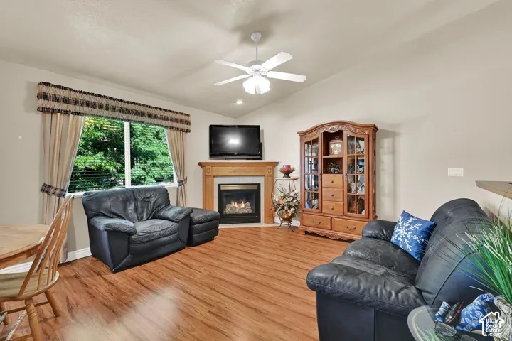 Vaulted ceiling with a ceiling fan, corner fireplace with a wood mantel, wood-finish flooring, and a large window