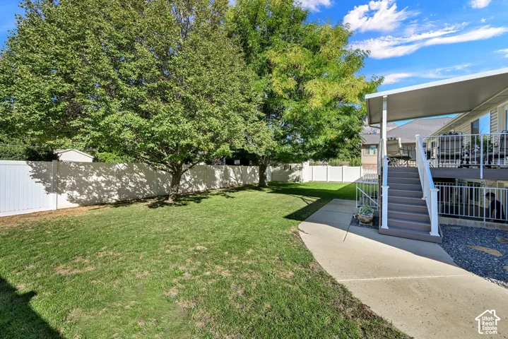 Expansive backyard with a manicured lawn, mature trees, white privacy fencing, a covered deck, and exterior stairs with white railings