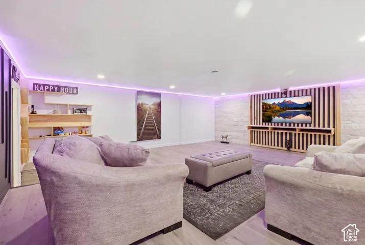 Luxurious recreation room featuring ambient purple LED ceiling lighting, plush upholstered seating throughout, built-in wooden shelving with display storage, wall-mounted entertainment display, and neutral wood-finish flooring with area rug accents