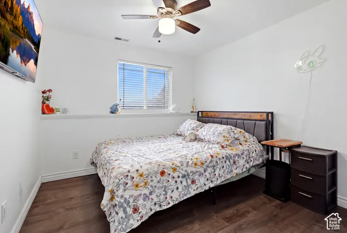 Bright secondary bedroom featuring dark wood-finish flooring, a ceiling fan with integrated lighting, and a large window with horizontal blinds providing natural light