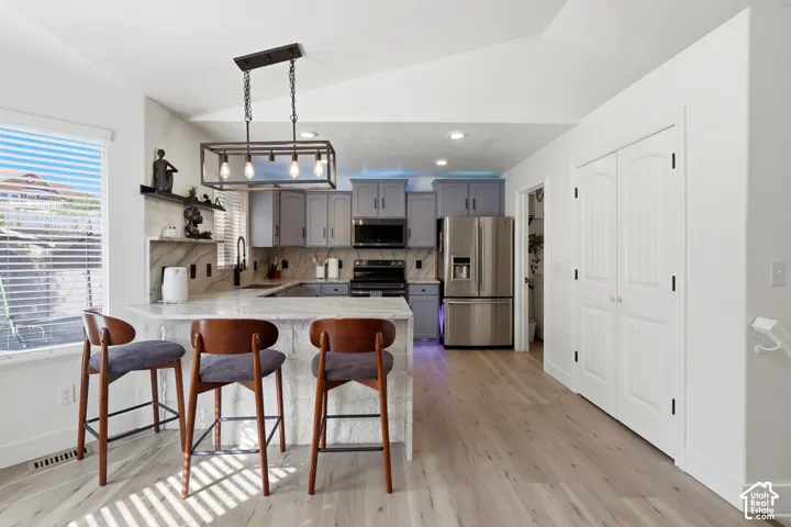 Kitchen featuring light wood-finish flooring, a large island with a counter-height bar, light gray cabinetry, stainless steel appliances, and a vaulted ceiling
