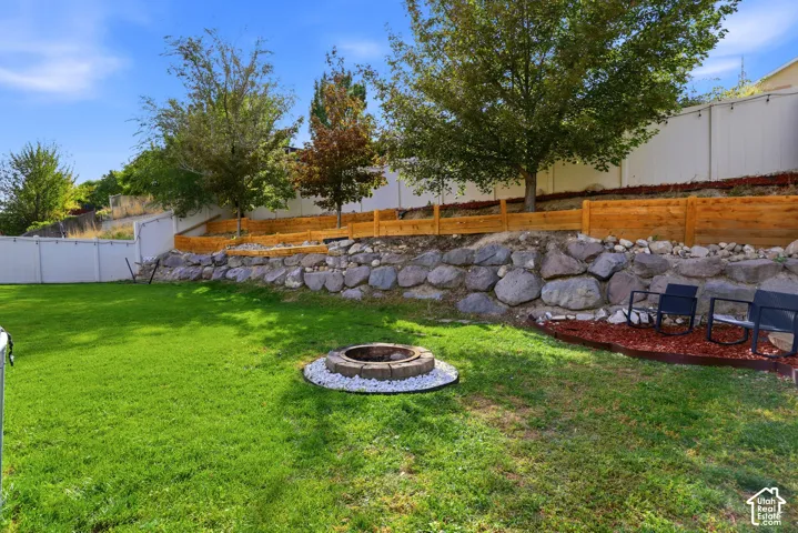 Expansive lawn area, built-in stone fire pit, tiered landscaping with a rock retaining wall, wood fencing, and mature trees