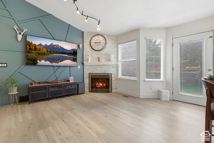 Wood-finish flooring, accent wall with geometric paneling, stone-clad fireplace with hearth, track lighting, and a glass-paneled exterior door