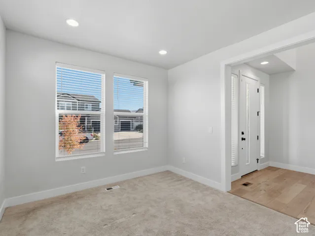 Recessed lighting illuminates the space, featuring two windows, light-toned carpeting, and a white entry door with a sidelight