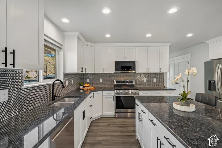 Kitchen featuring white shaker cabinetry, dark granite countertops, stainless steel appliances, a black penny tile backsplash, and wood-finish flooring