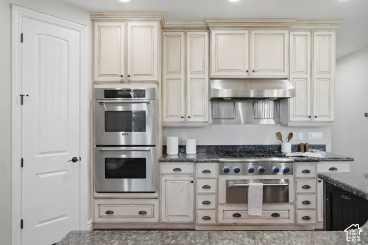 Custom cabinetry with a distressed finish, stone countertops, stainless steel double wall ovens, stainless steel gas range, and stainless steel range hood