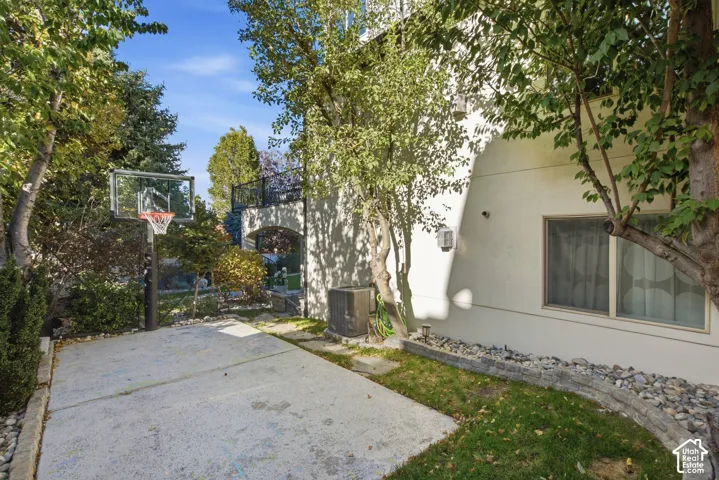 Concrete sport court featuring a basketball hoop, stucco exterior finish, elevated balcony with metal railing, and stone-accented landscaping