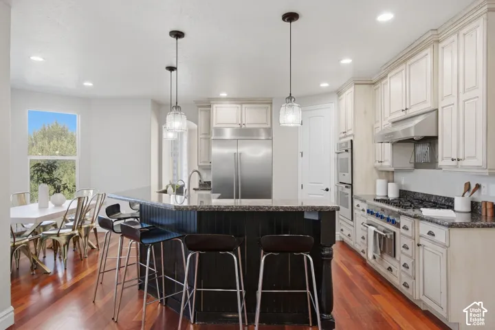 Kitchen featuring wood-finish flooring, custom cabinetry with a contrasting island, stone-surfaced countertops, stainless steel appliances, and pendant lighting