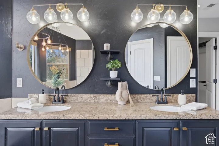 Double vanity featuring navy cabinetry, gold-tone hardware, a stone-composite countertop, dual undermount sinks, and round mirrors with gold-tone frames